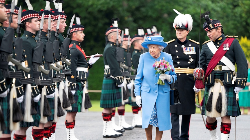 Queen Elizabeth dressed in blue surrounded by people dressed in traditional scottish uniforms with quilts