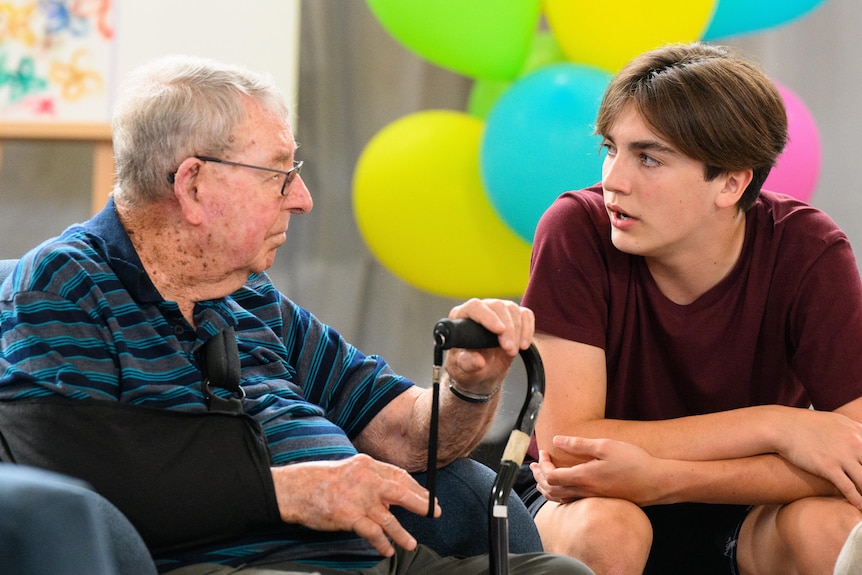 An elderly man in a blue shirt sits in an armchair across from a teenage boy in a maroon shirt.