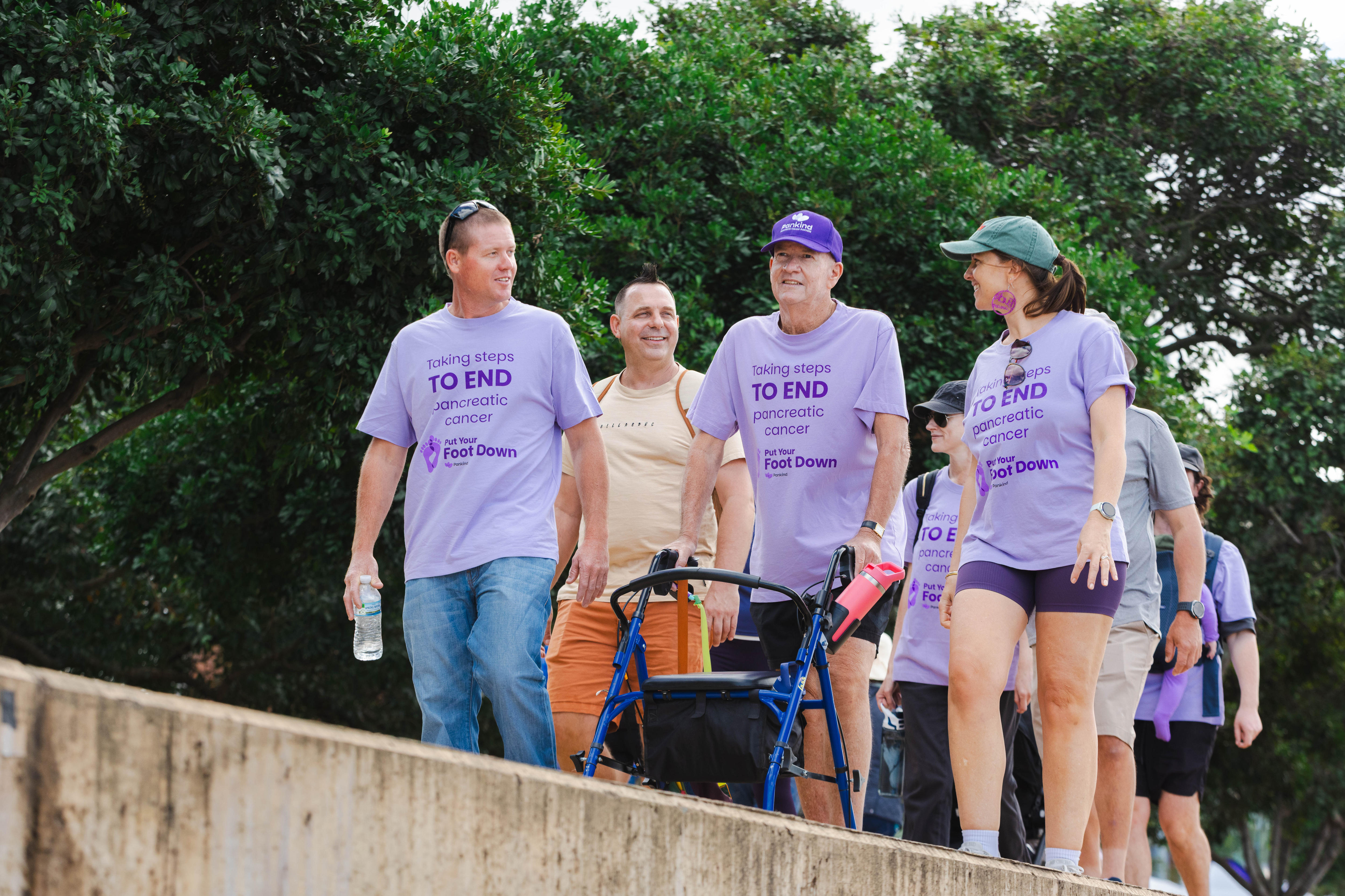 A man in a purple shirt and hat pushing a walker walks along a bridge with a man on one side and a woman on the other 