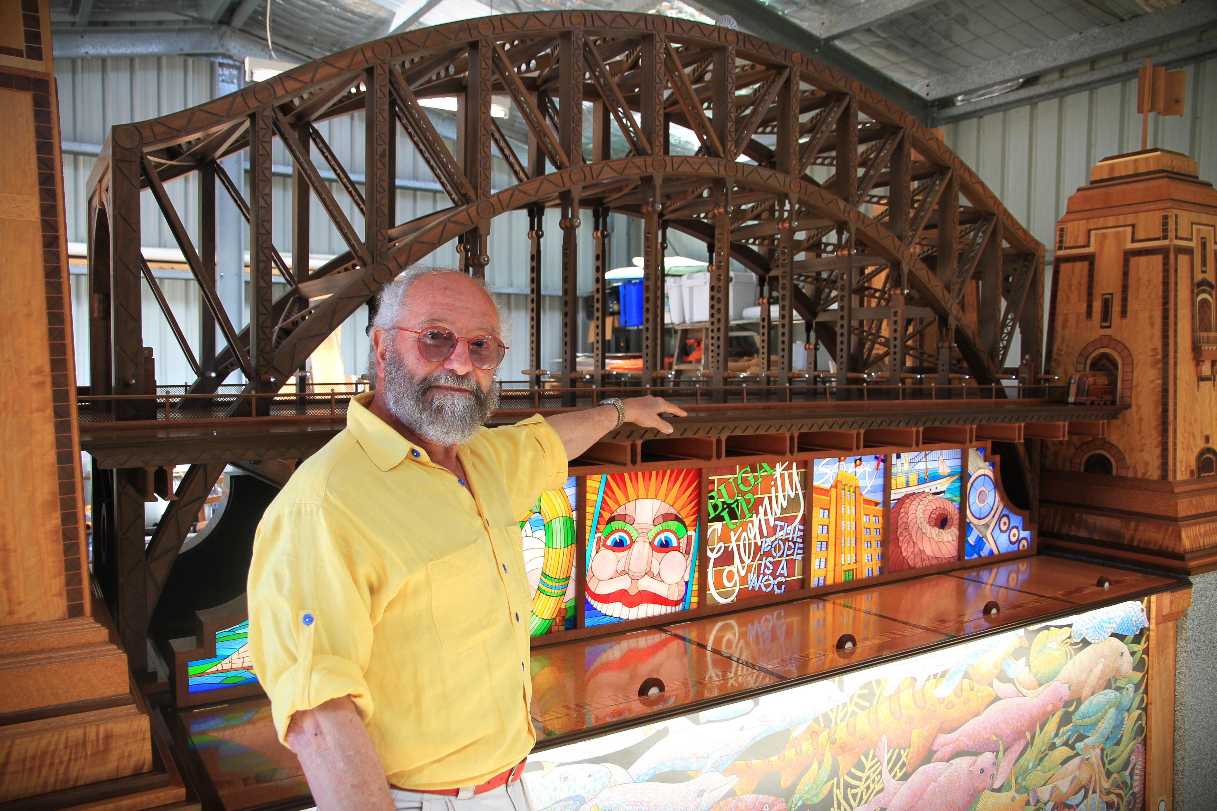 Men with grey hair, glasses and yellow shirt, holding arm on miniature Sydney Harbour Bridge