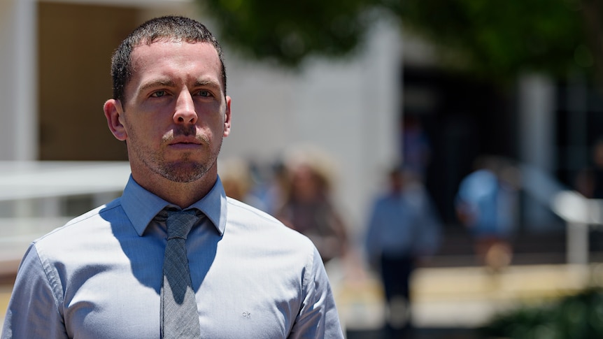 A man in a collared shirt and tie looking serious, standing outside a court building.