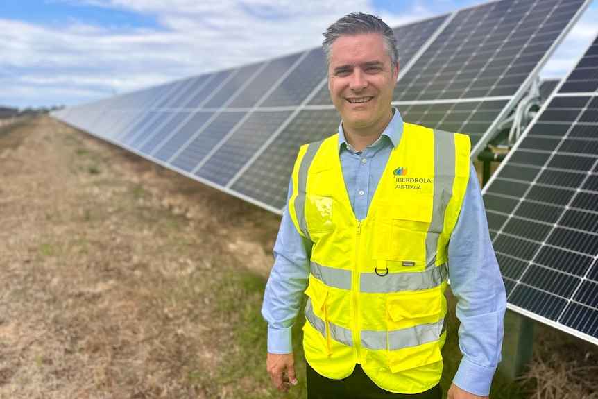 Man in a blue business shirt and yellow hi-vis vest standing in front of a row of solar panels.