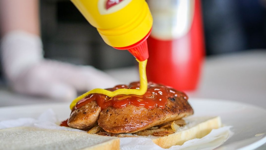 A close-up shot of a bottle of mustard being squeezed onto a butterflied sausage on bread and onions, with tomato sauce on top.