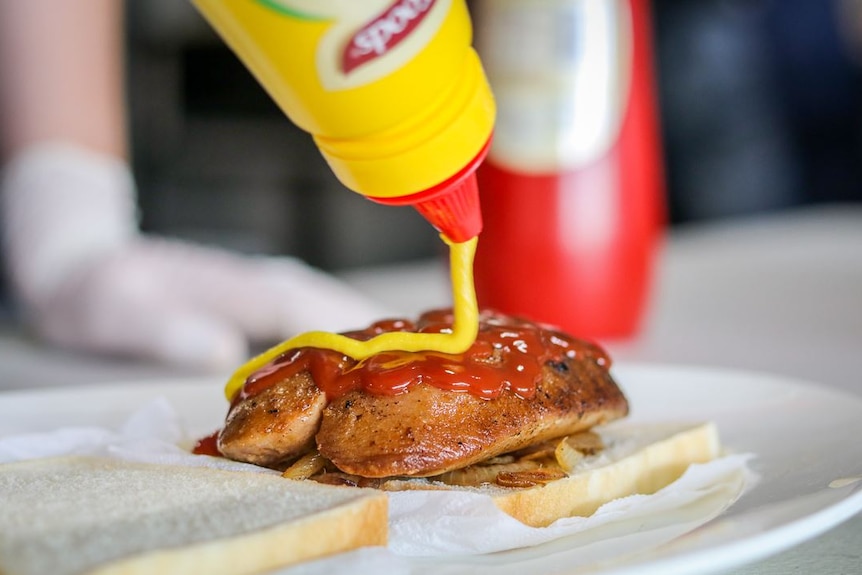 A close-up shot of a bottle of mustard being squeezed onto a butterflied sausage on bread and onions, with tomato sauce on top.