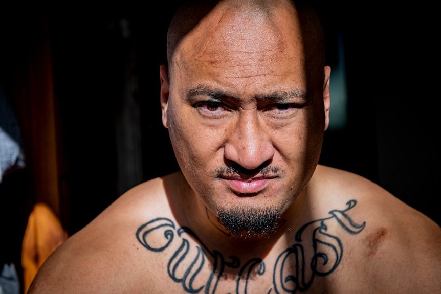 A man stares at the camera, he is shirtless, with the words "Outcast" tattooed on his chest.