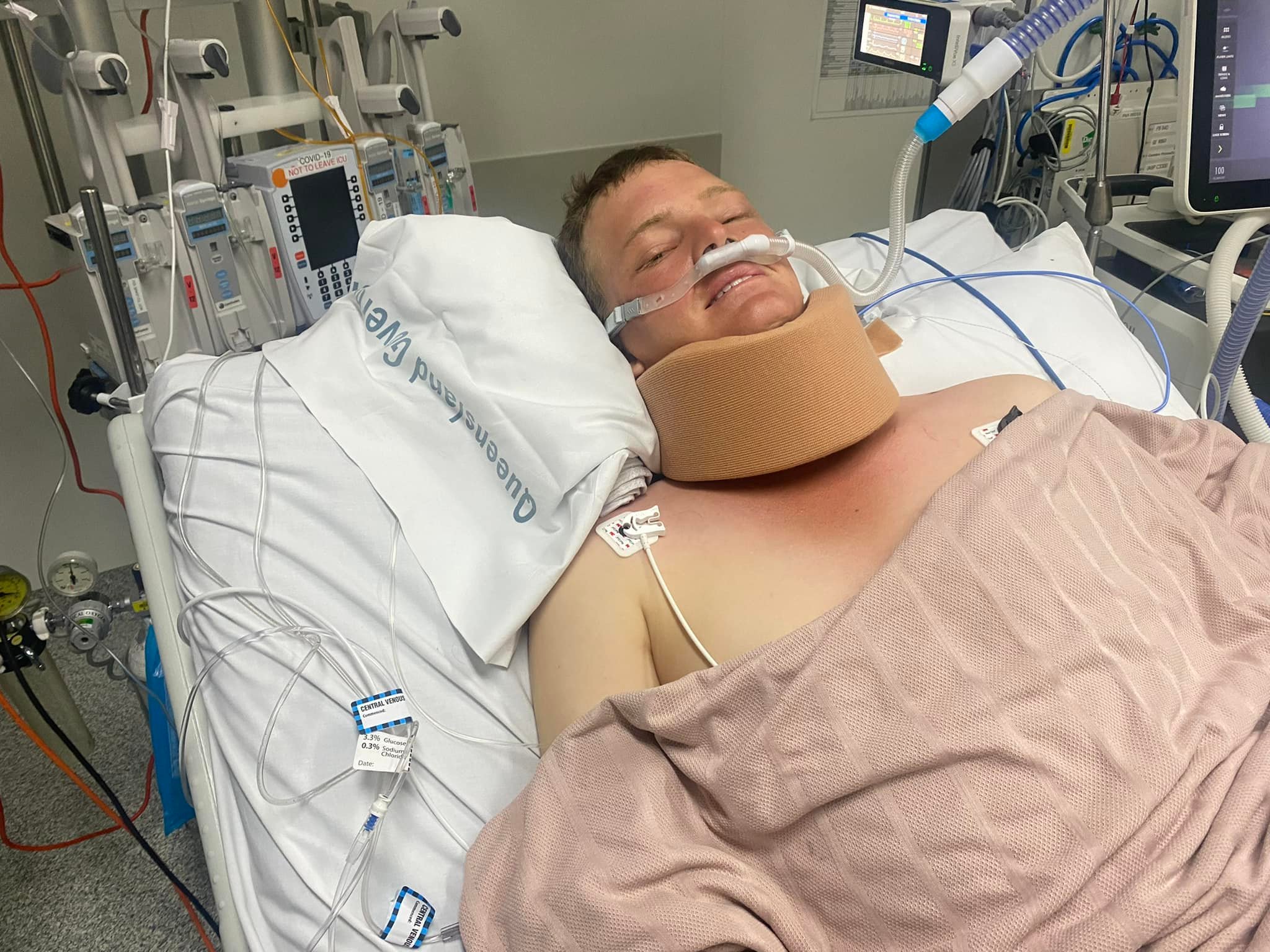 A young man in hospital bed appears to smile despite a neck brace, oxygen and tubes. 