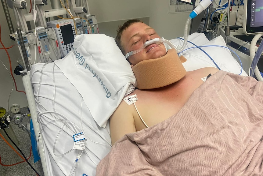 A young man in hospital bed appears to smile despite a neck brace, oxygen and tubes. 