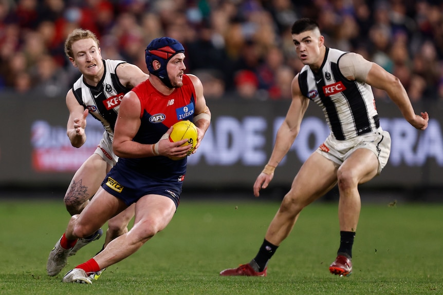 A Melbourne AFL player wheels to his left with the ball as Collingwood defenders adjust to try to stop him.