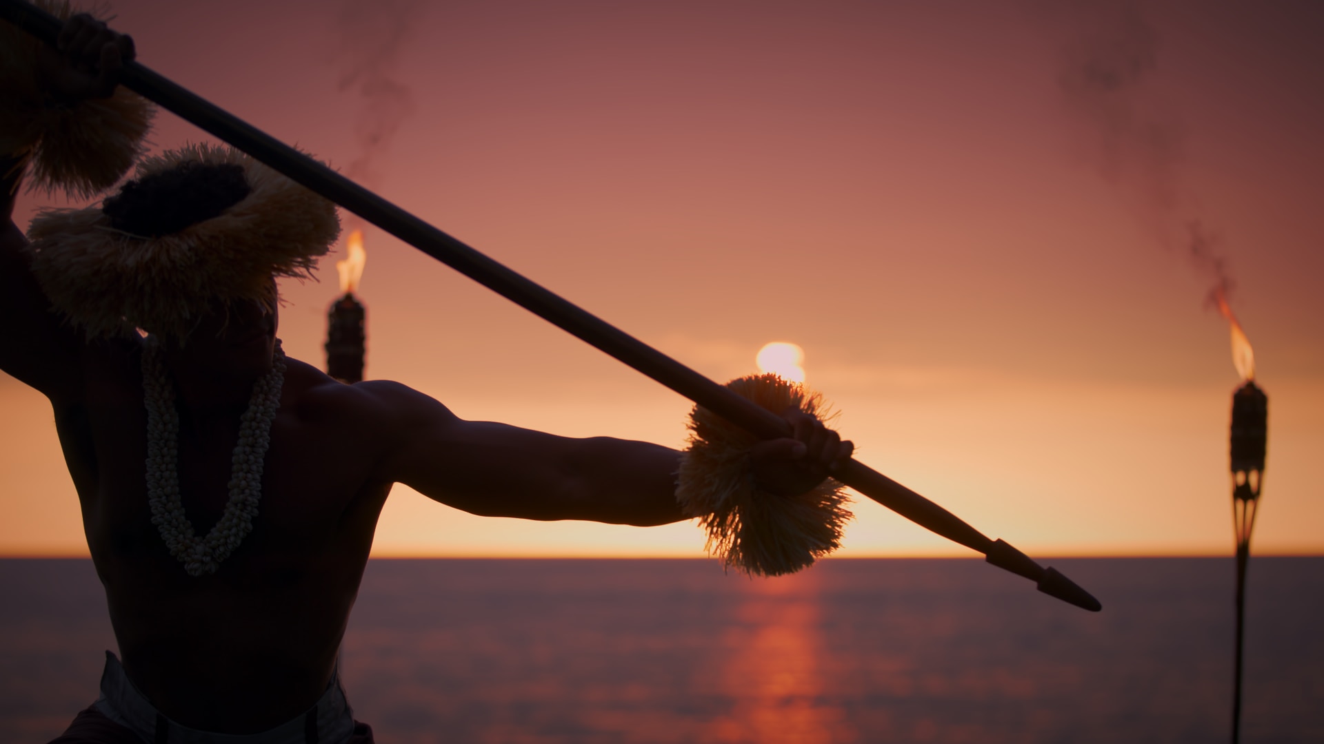 A man bathed in shadow holds a spear up at sunset in front of a beach while performing.