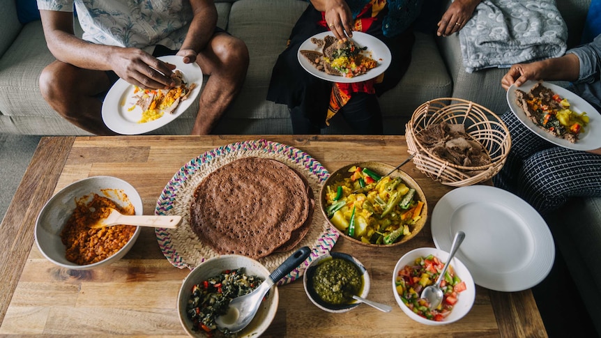 Plates and bowls of Ethiopian food on a coffee table with three people sitting on a sofa around it and eating.
