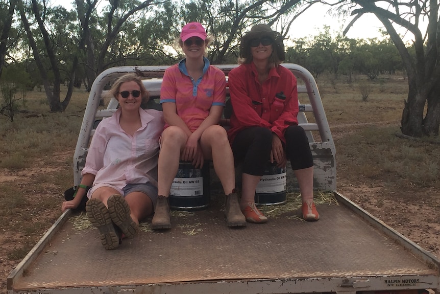 Three women sit on the back of a ute, dressed in colours of pink, and red, trees behind them. 
