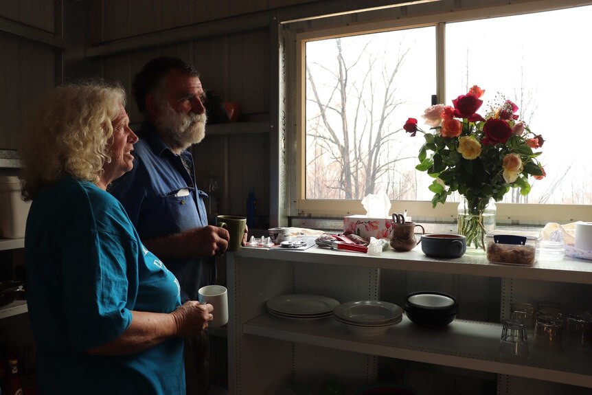 A man and a woman hold mugs of tea and look out the window in a kitchenette area.