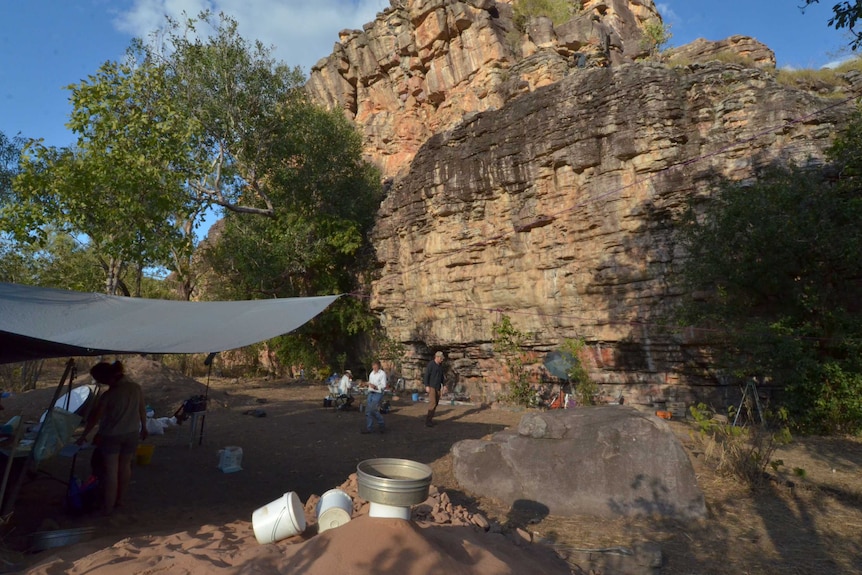 A large rock wall, with a group of people doing excavation work below it