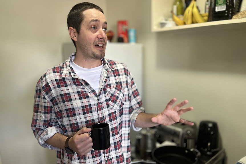 A man wearing a checked shirt in a kitchen at holding a mug looking off camera