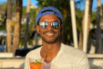Tanila De Silva, a young man wearing sunglasses and a cap, smiles happily on a beach.