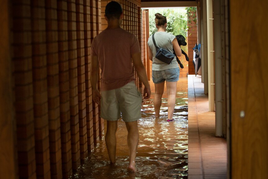 Man and woman walk through puddles in house holding a dog. 