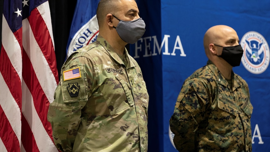 Two US military personnel stand wearing masks in front of an American flag and FEMA banner. 