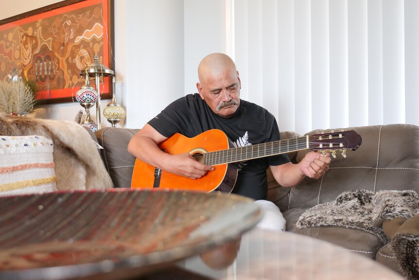A Maori man tunes an acoustic guitar while sitting on a couch inside a living room 