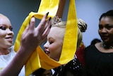 A woman with albinism is crowned with a plastic tiara and a bright yellow sash in a dimly-lit room.