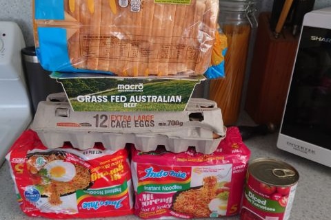 bread, beef mince, eggs, noodles and a can of diced tomatoes stacked on a bench.