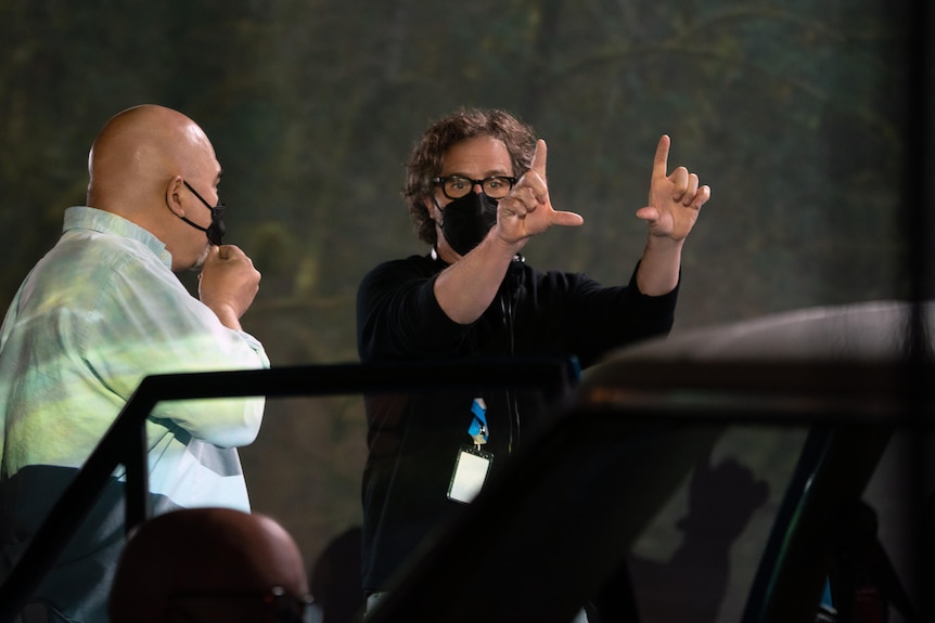 A man with short curly hair, wearing a black face mask, makes a gesture with his hands similar to a photo frame.