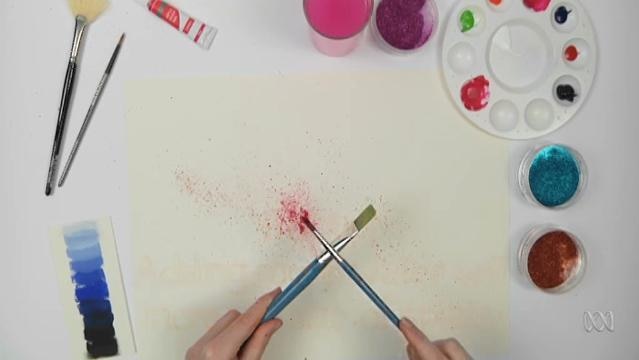 Hand uses two paintbrushes to make splatter patterns on paper