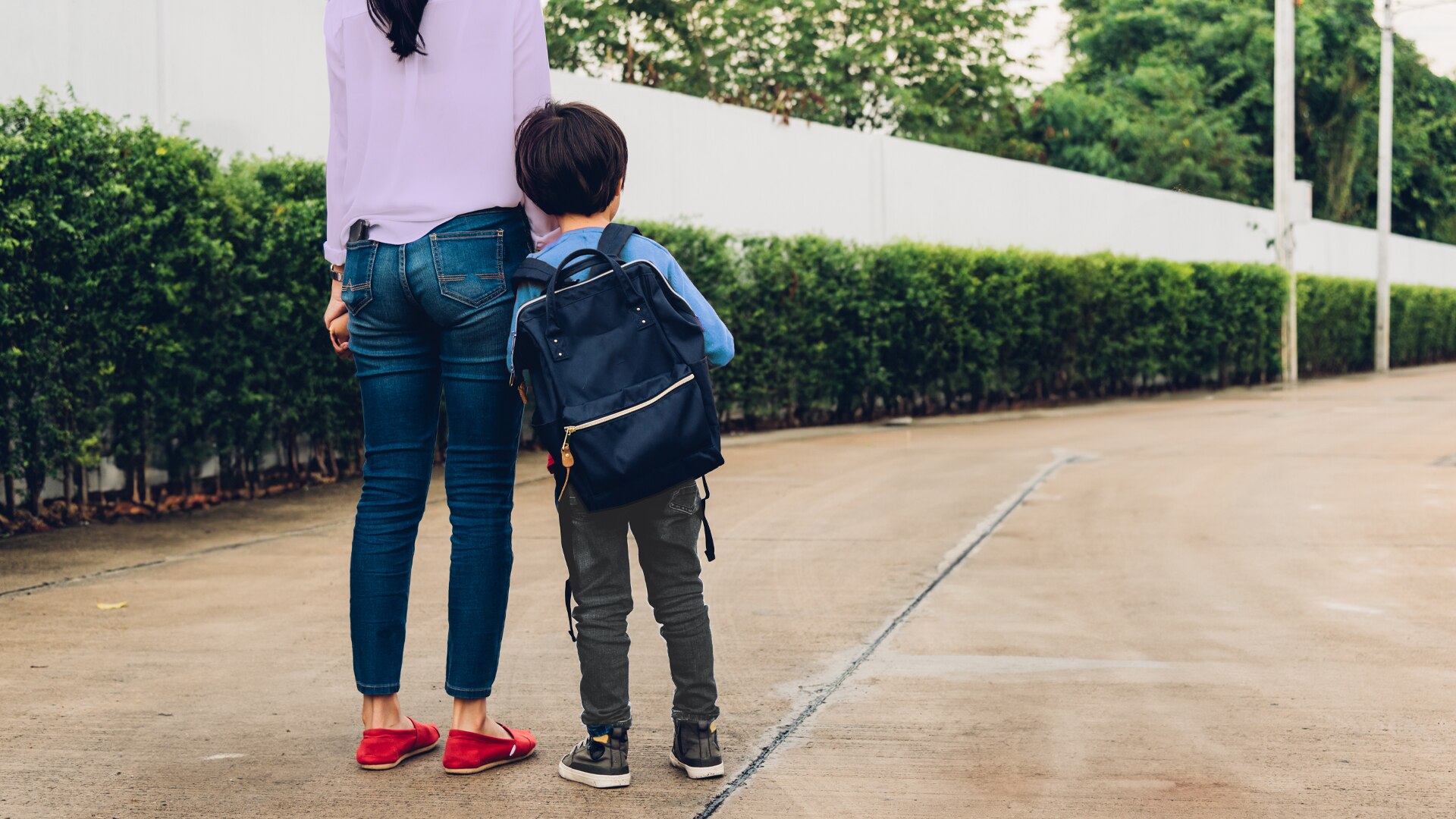 A woman holds the hand of a young boy wearing a school backpack.