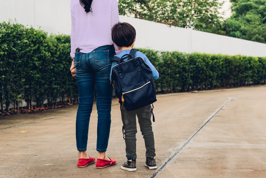 A woman holds the hand of a young boy wearing a school backpack.