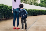 A woman holds the hand of a young boy wearing a school backpack.