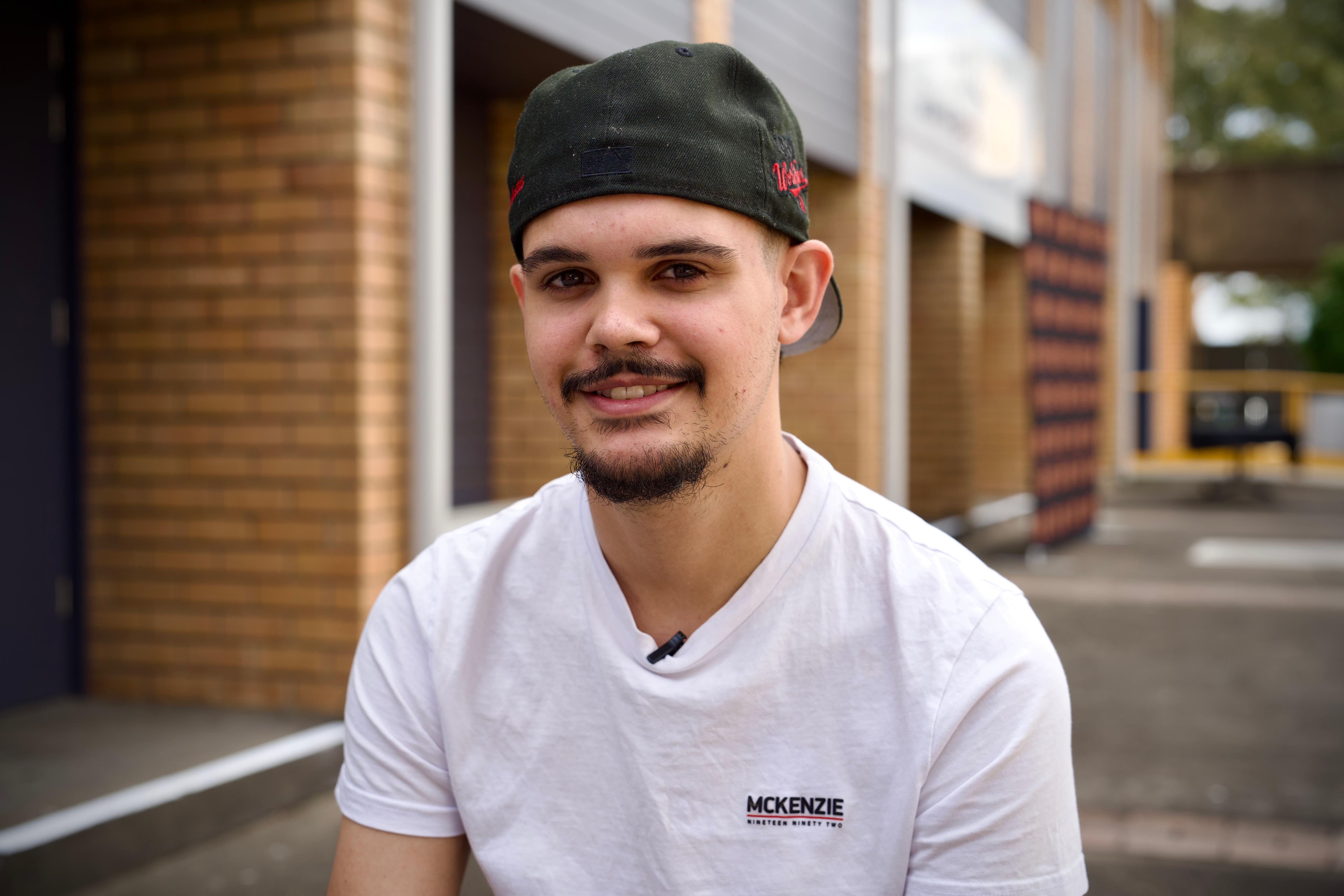 A young Indigenous man sitting outside smiles at the camera wearing a white t-shirt and a black backwards cap