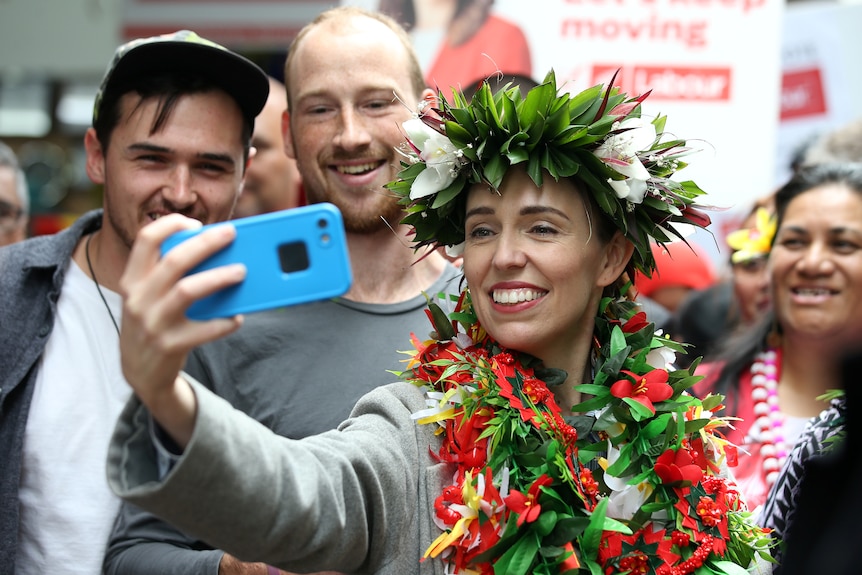 Jacinda Ardern, wearing a floral head piece and necklace, smiles while taking a selfie on the campaign trail