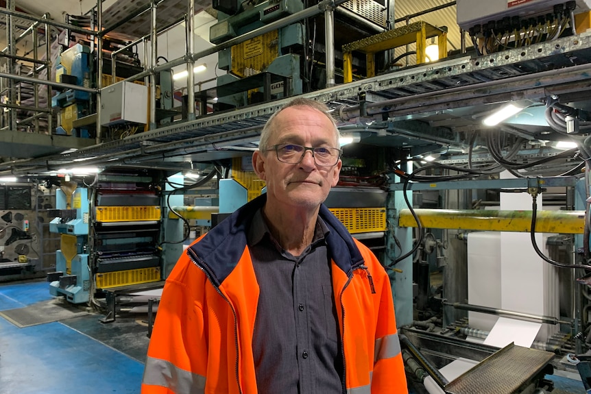 A man with a blue shirt, glasses and high-visibility jacket stands in front of an industrial printing press.