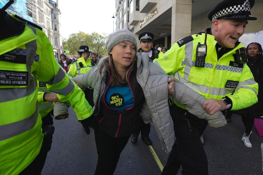 Greta Thunberg in a beanie and wearing a button reading "Oily Money Out" is taken away by police in hi vis uniforms