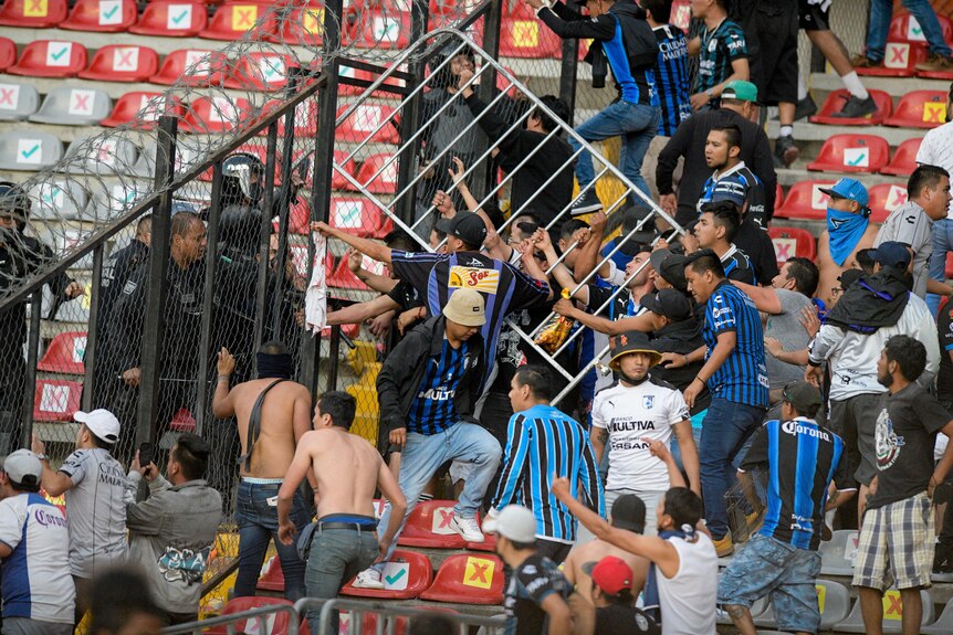 Men gather in the stands, facing security at a fence. Some holding a metal barrier 