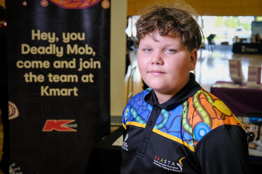 An Indigenous boy standing next to a sign promoting jobs for Indigenous students at Kmart
