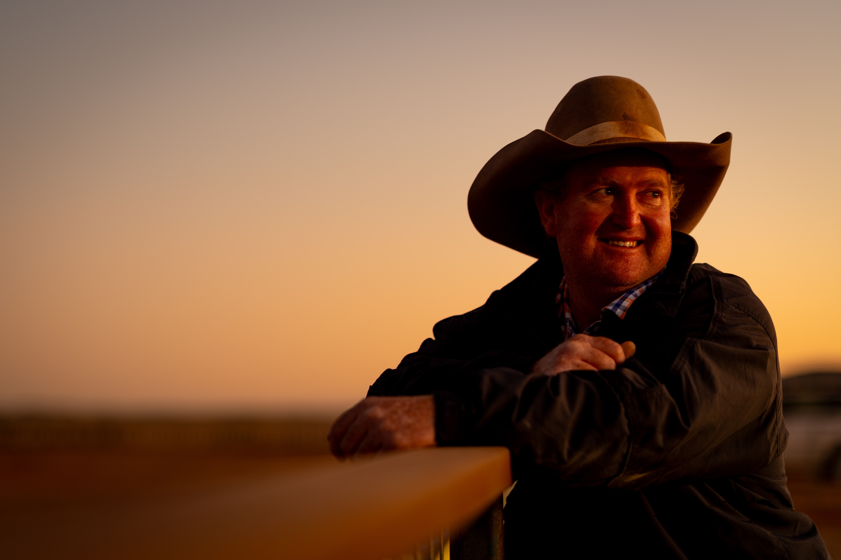 A man wearing a cowboy hat smiling to the right of screen, leaning against a fence