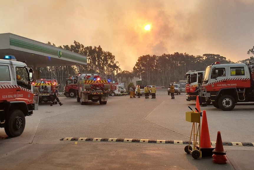 fire fighters at parking lot with black smoke rising behind the trees in the background