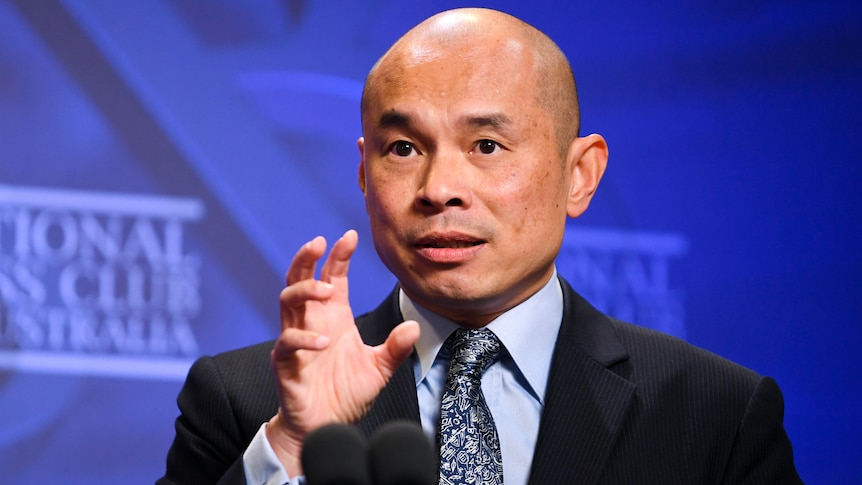 A man in a suit and tie holds his right hand up as he speaks into a microphone.