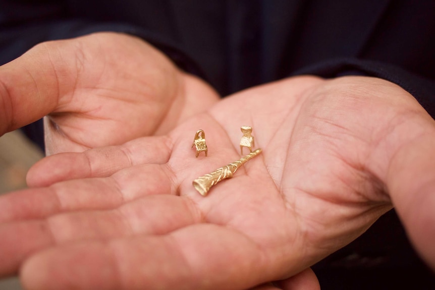 Close up of Sameer Dakhil's hands. He is holding three brass miniature figures: two chairs and a didgeridoo.