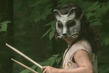 A young girl in a flower print dress and an unsettling cat mask plays a drum that is suspended around her neck.