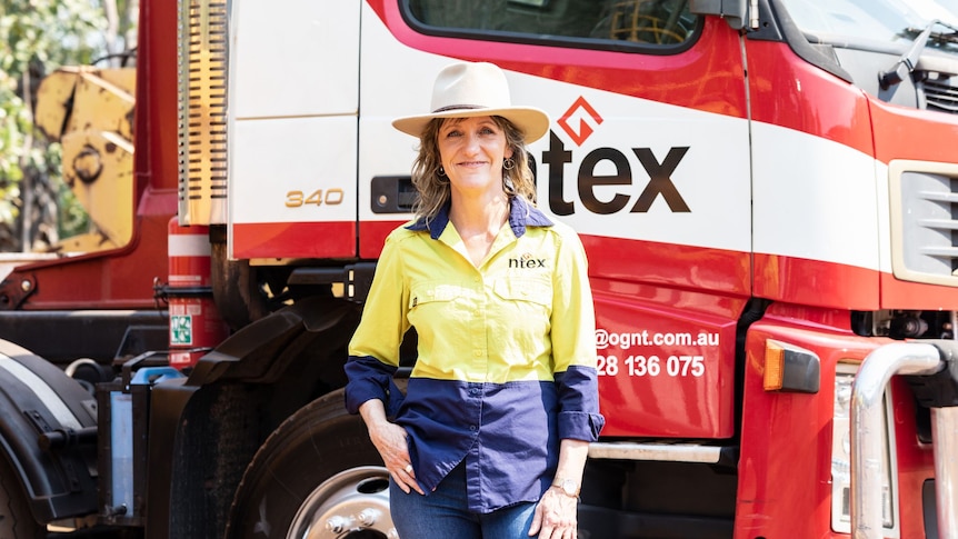A woman wearing an akubra hat and hi-viz shirt stands proudly in front of a truck.