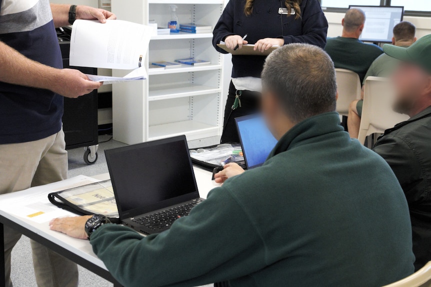 inside a classroom at a jail. there are two inmates sitting infront of laptops. Their faces are blurred