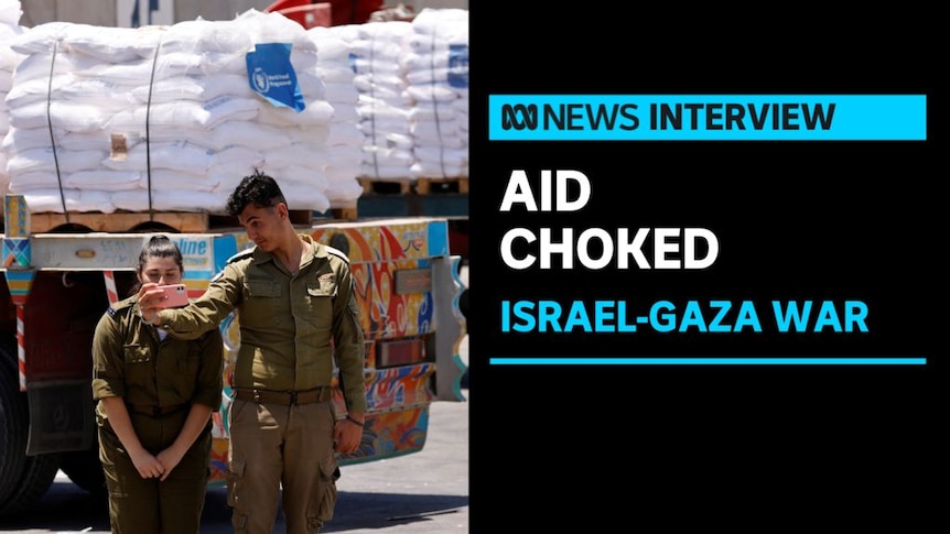 Aid Choked, Israel-Gaza War: Two people in military fatigues stand next to each other with aid on trucks in the background.
