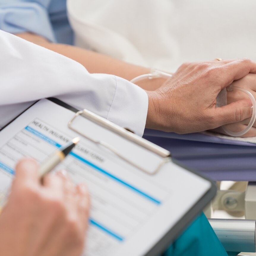 Close up of clipboard in foreground, and doctor's hand resting on hand of patient in hospital bed.