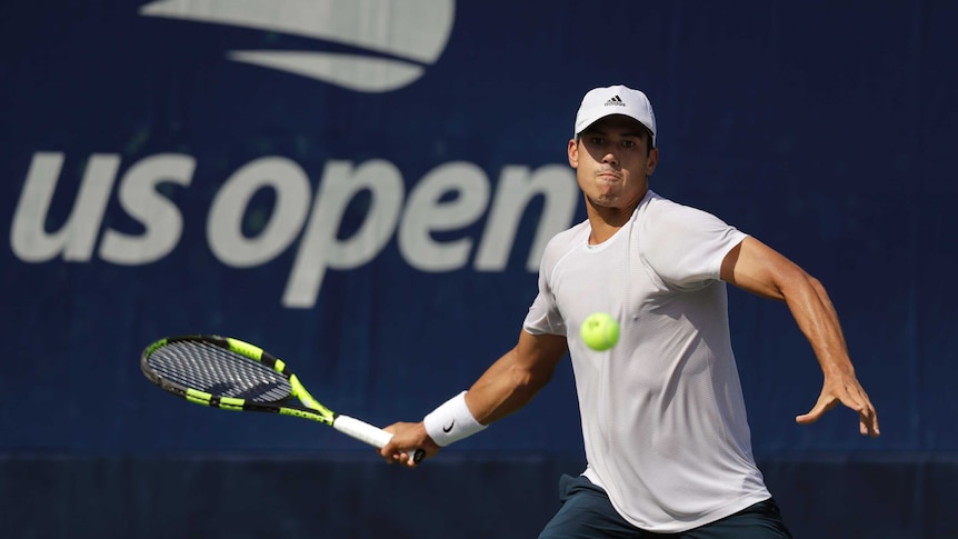 A male tennis player wearing a grey shirt, blue shorts and a white hat, watches the ball.