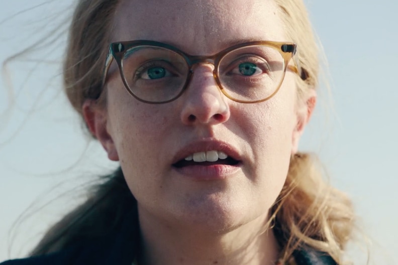 Blue-eyed bespectacled woman's pulled back blonde hair has blank expression on clear sky day, her face half lit by the sun.