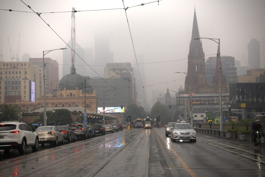 Cars drive over Princes Bridge in Melbourne towards Flinders Street Station and the CBD which are shrouded in a grey haze.