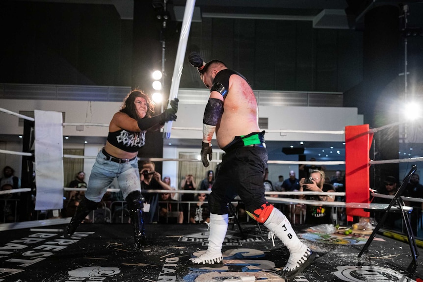 In a boxing ring, a woman swings a long light globe down towards the head of a man who attempts to block it with a fist.