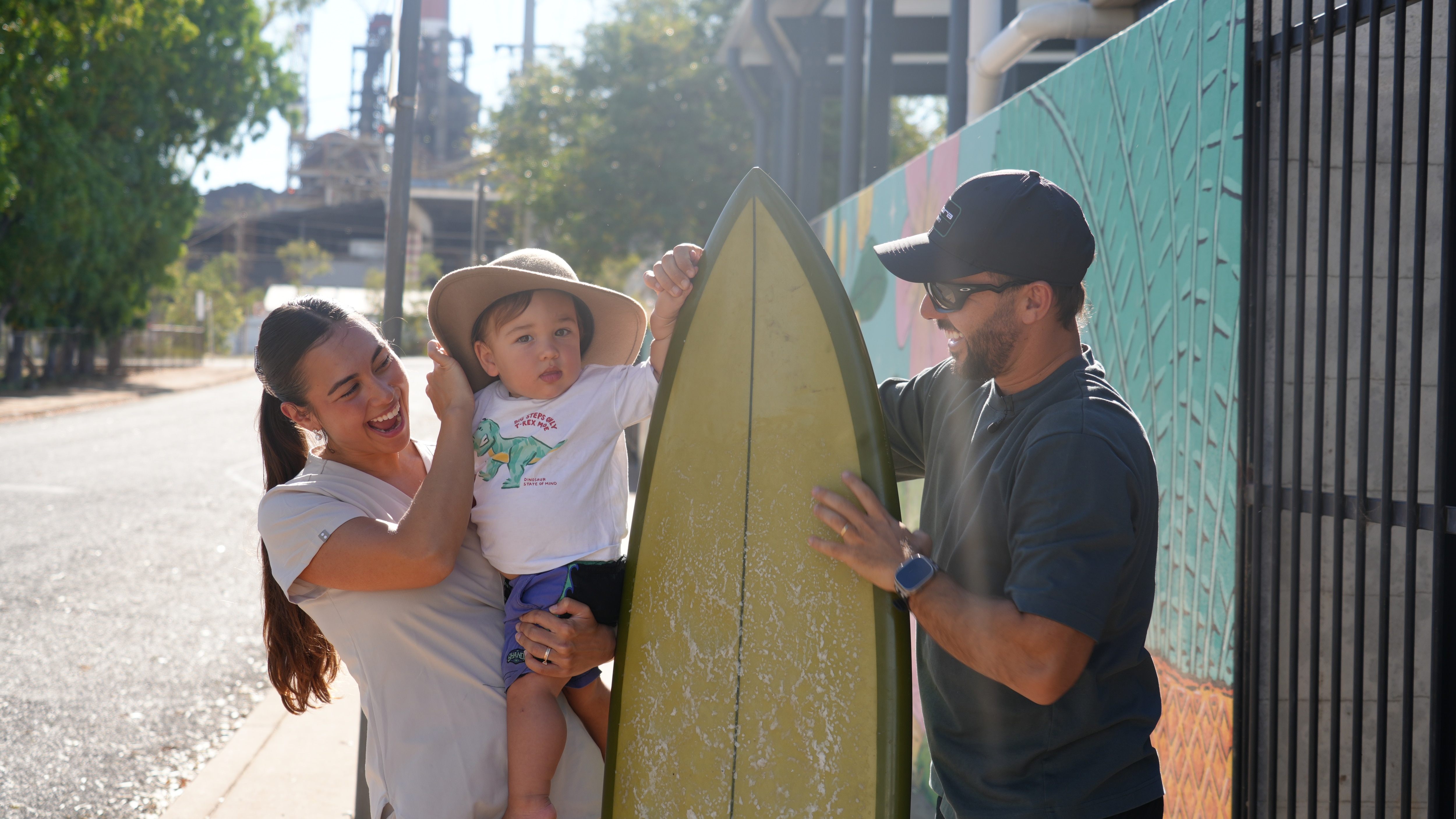 a wife, husband, and toddler standing in a suburban street smiling with a surboard between them