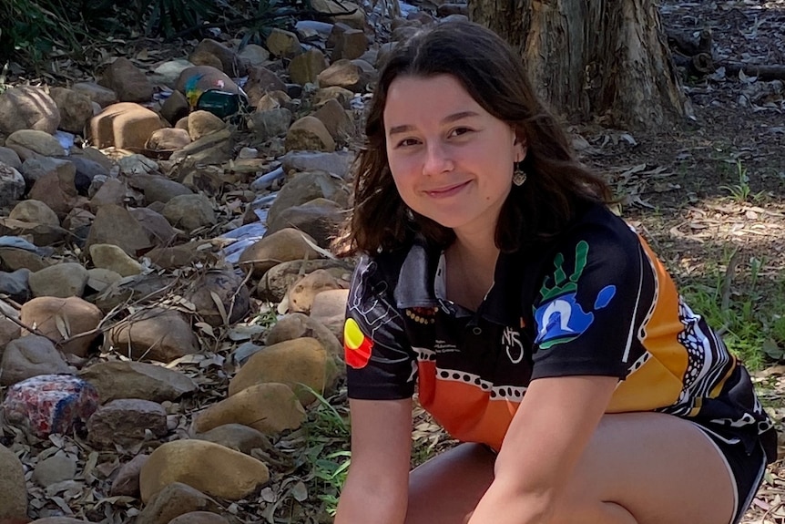 Zali Reynolds crouches on the ground outdoors, smiling at the camera while painting a small rock.